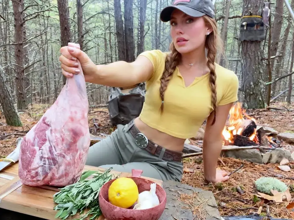 Out in a pine forest, a woman in a cap shows off a raw leg of lamb, with herbs, a lemon and garlic in front of her and a fire going in the background